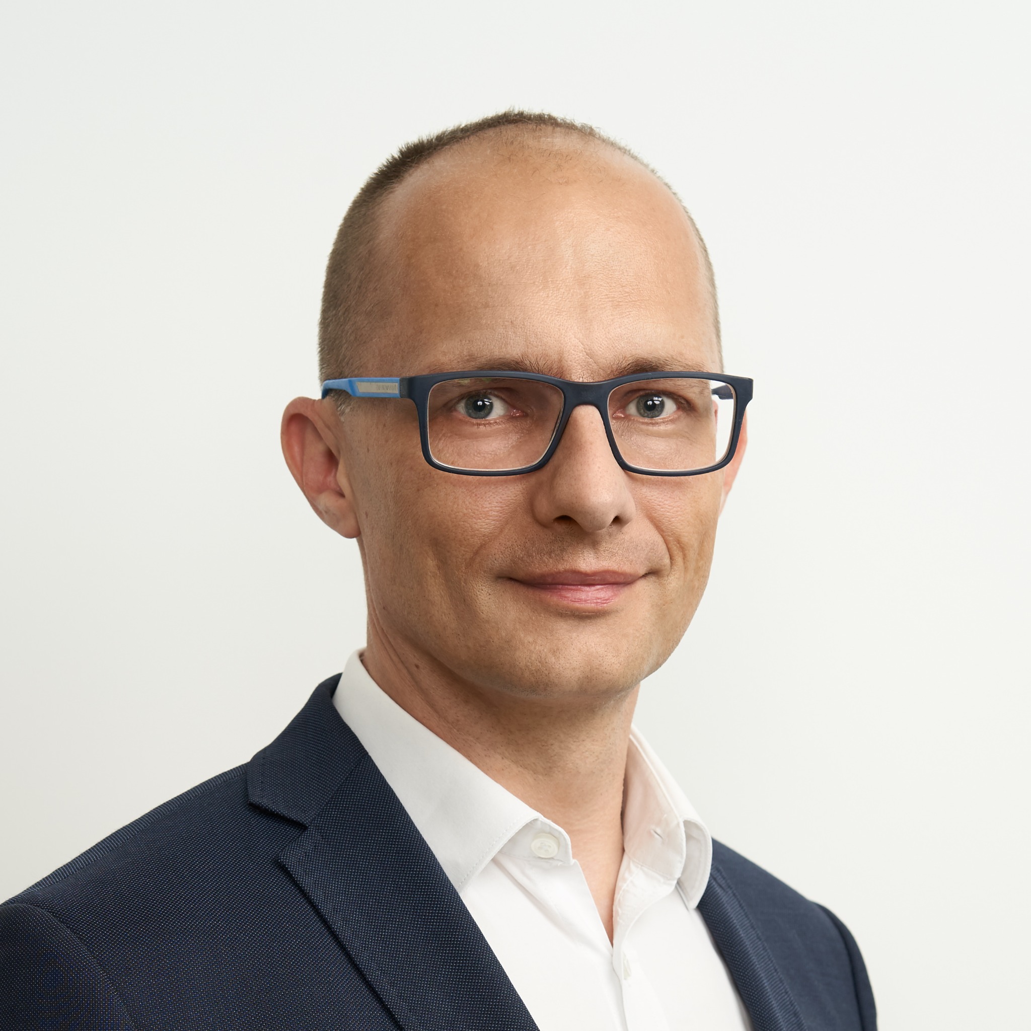 Portret mężczyzny w garniturze i okularach z niebieskimi zausznikami, uśmiechającego się do kamery na jasnym tle. Profesjonalny wizerunek doradcy.