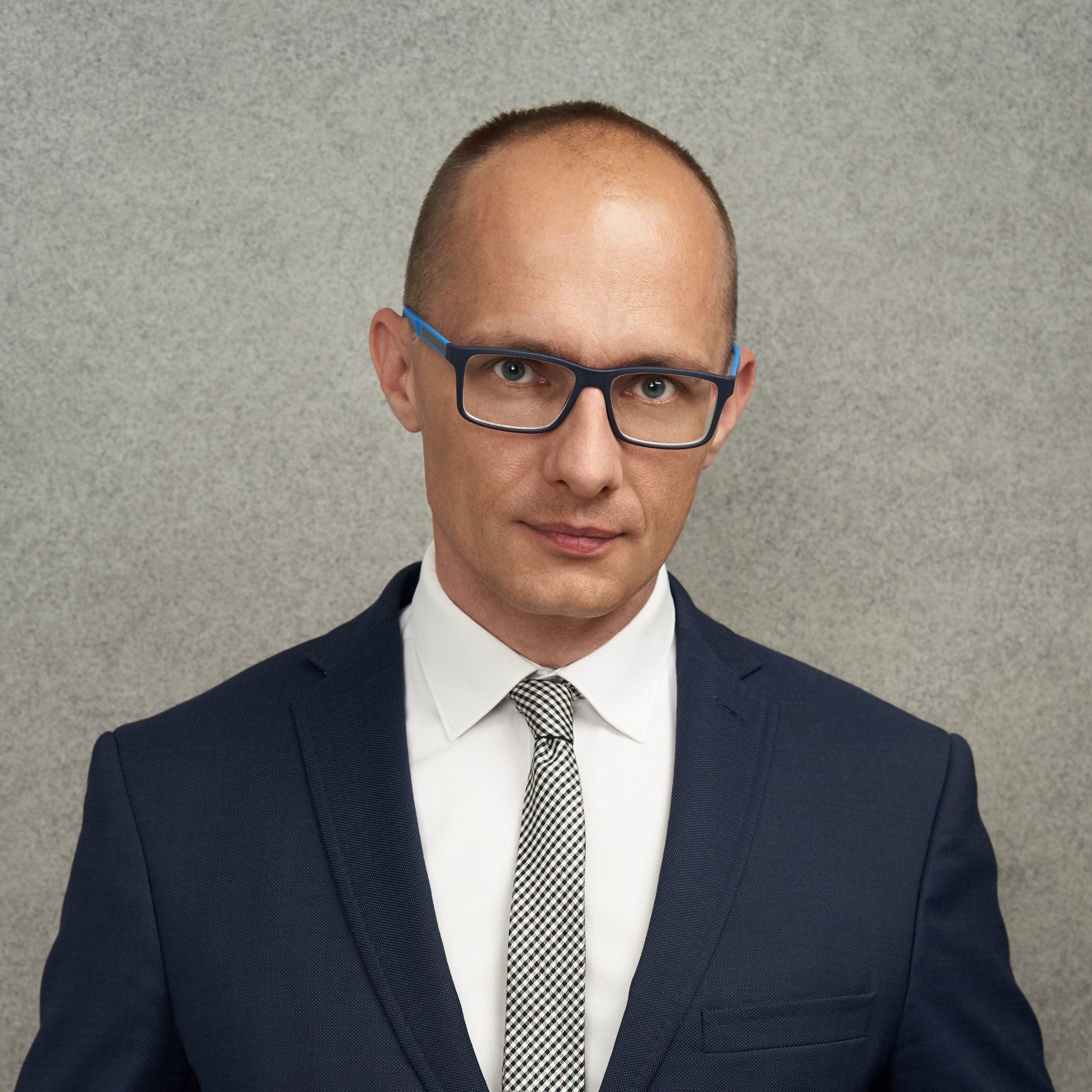 Portret mężczyzny w garniturze i okularach z niebieskimi zausznikami na tle szarej ściany. Uśmiech na twarzy, wzrok skierowany wprost. Profesjonalny wizerunek.