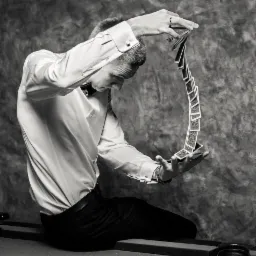 Czarno-białe zdjęcie iluzjonisty w białej koszuli i czarnych spodniach, siedzącego i prezentującego trik z kartami unoszącymi się w powietrzu między jego dłońmi na tle szarej tekstury.