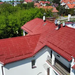 Pracownik w kamizelce na dachu z czerwonej blachodachówki, montujący antenę satelitarną, widok z góry na dom w zabudowie szeregowej i okoliczną zieleń.