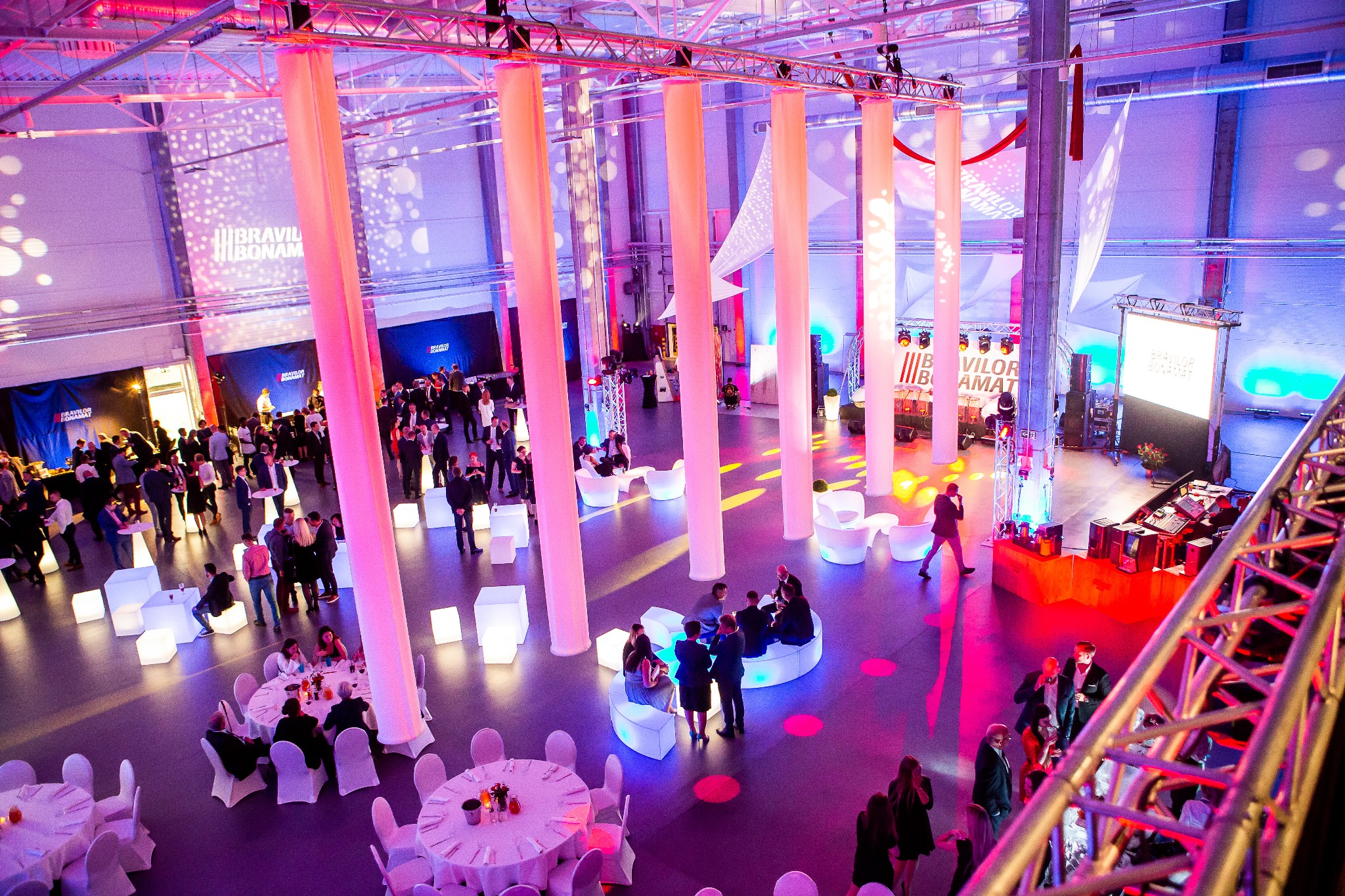 Szeroki plan sali konferencyjnej oświetlonej różowymi i fioletowymi światłami, z grupami ludzi siedzącymi przy okrągłych stołach i na białych, podświetlanych kostkach, widok z góry z elementami...