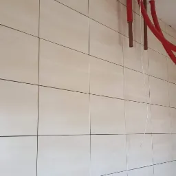 Ściana wyłożona jasnymi, prostokątnymi płytkami ceramicznymi, z widocznymi przewodami instalacji elektrycznej i rurami centralnego ogrzewania wystającymi ze ściany i sufitu, częściowo...