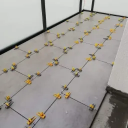 Układanie szarych płytek podłogowych z użyciem klipsów i klinów poziomujących, widoczna fuga i fragment ściany z oknem.