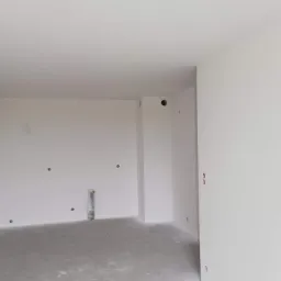 Wnętrze pokoju po szpachlowaniu, ściany i sufit pokryte białą gładzią, widoczne punkty elektryczne i pion kanalizacyjny zabezpieczony folią.