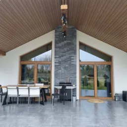 Zadaszony taras z drewnianym sufitem, dużymi oknami z brązowymi ramami i szarą kamienną ścianą pomiędzy nimi, na szarej podłodze stoi stół z krzesłami i grill.