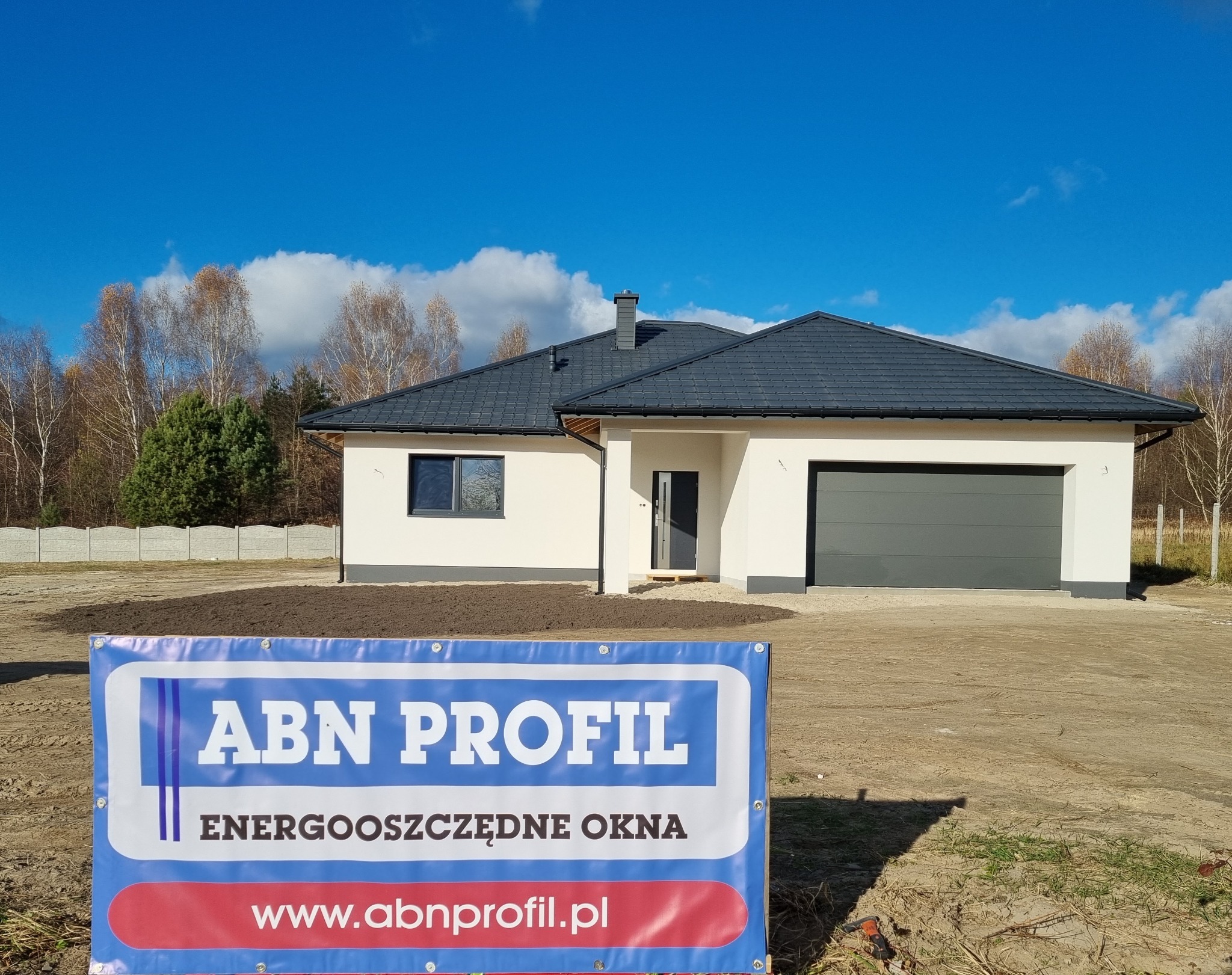 Nowoczesny dom jednorodzinny z szarą stolarką okienną i garażową, prezentowany z banerem reklamowym firmy ABN Profil na pierwszym planie.