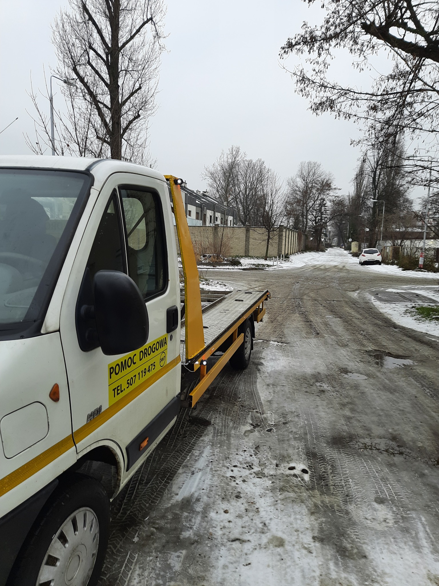 Biała laweta z żółtym napisem 'POMOC DROGOWA' i numerem telefonu, zaparkowana na ośnieżonej drodze w pochmurny dzień. Widoczny fragment kabiny i platforma załadunkowa.