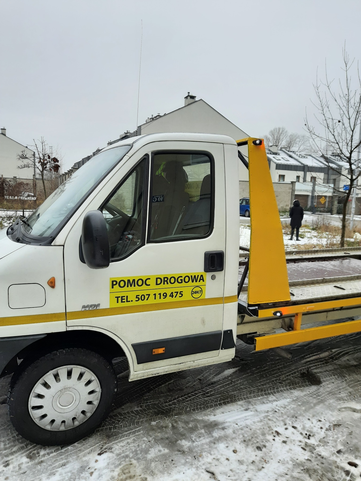 Biała laweta z żółtym wysięgnikiem i napisem 'Pomoc Drogowa' na tle zimowego krajobrazu miejskiego, widoczne ślady opon na śniegu.