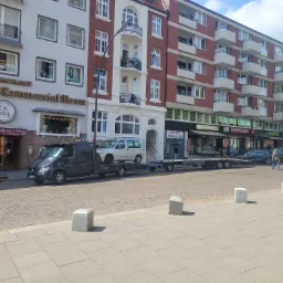 Czarna laweta z platformą transportową przewozi srebrnego vana na tle miejskiej zabudowy z restauracjami i apartamentami w Hamburgu.