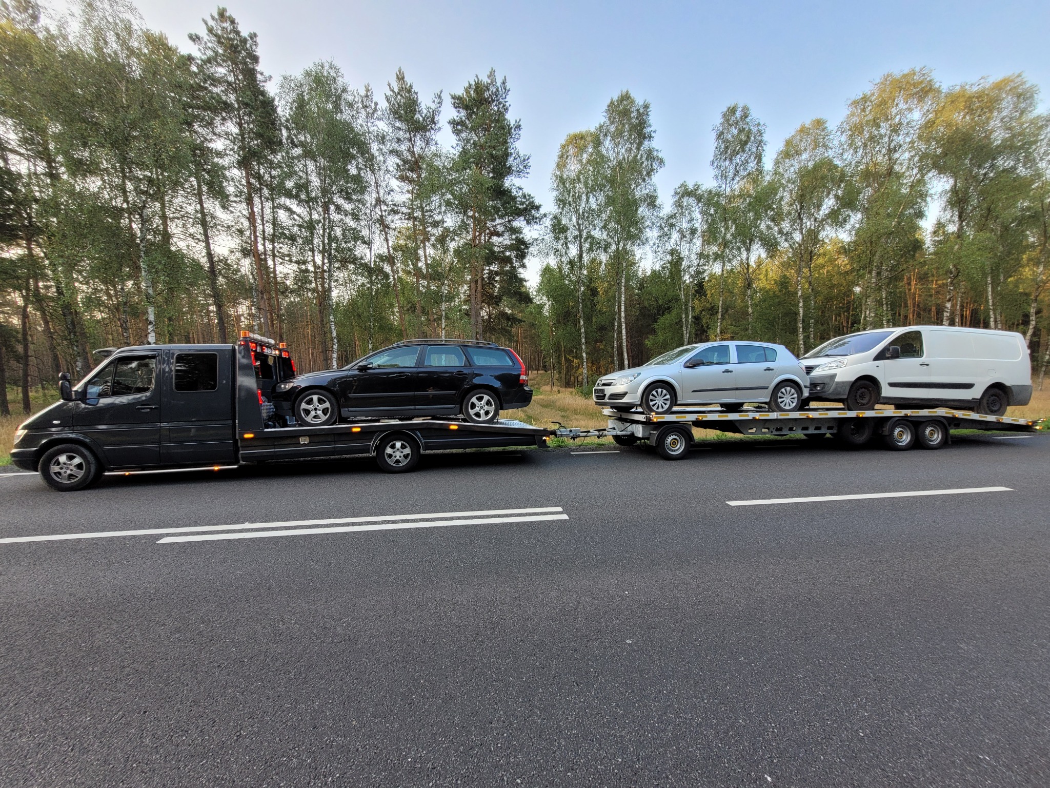 Czarna laweta z dwoma przyczepami przewożąca czarne kombi, srebrny hatchback i biały van na tle lasu.