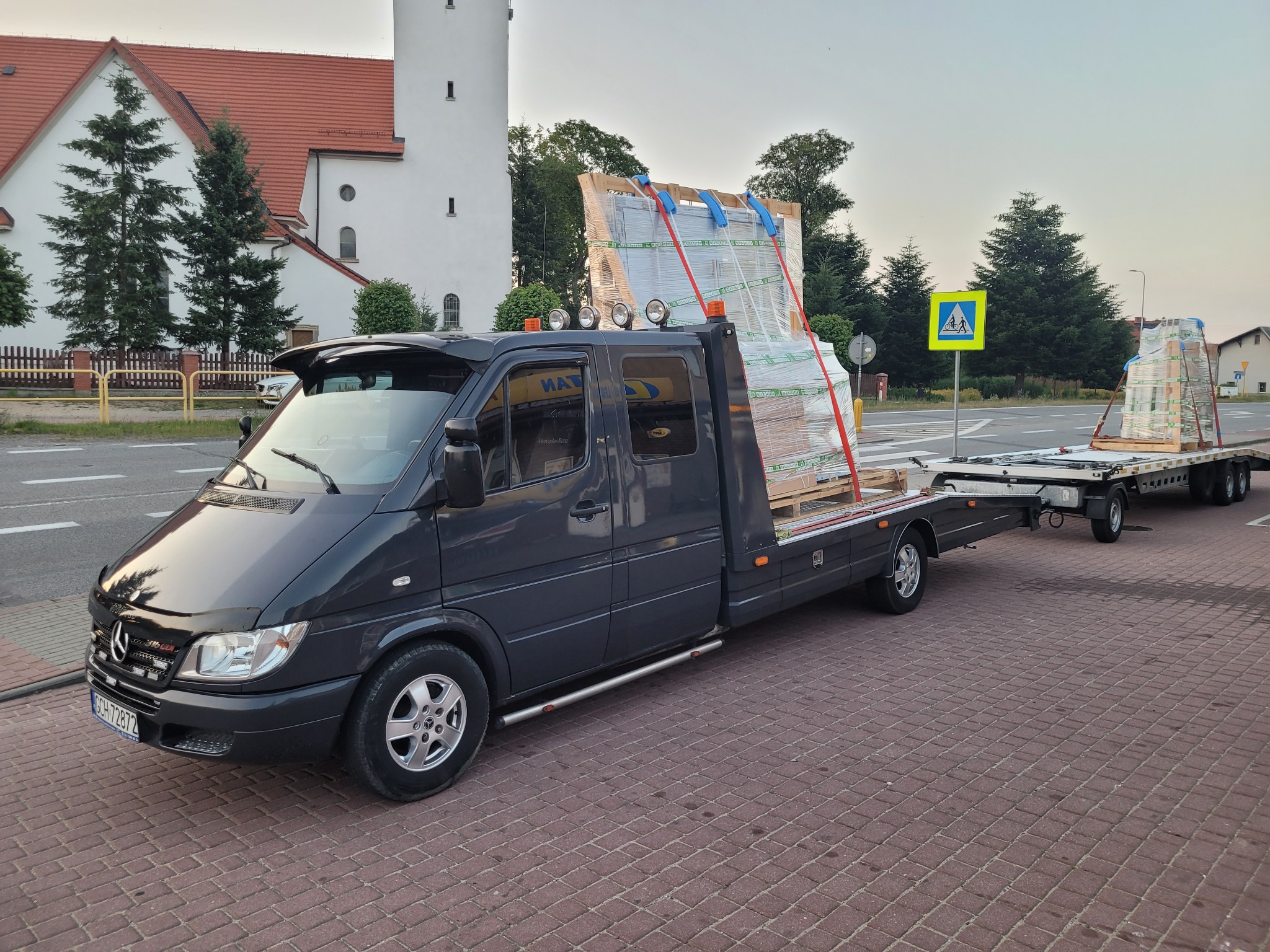 Czarny bus z lawetą przewozi zabezpieczony ładunek w pobliżu kościoła i znaku przejścia dla pieszych.