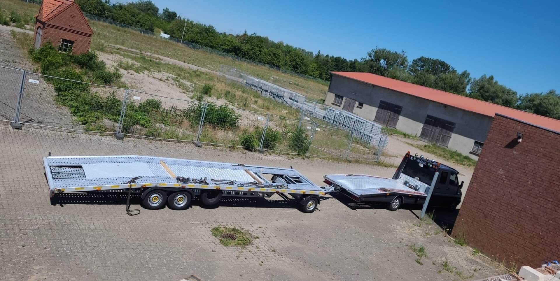 Czarna laweta z długą, srebrną platformą do transportu pojazdów, zaparkowana na placu z budynkami w tle.