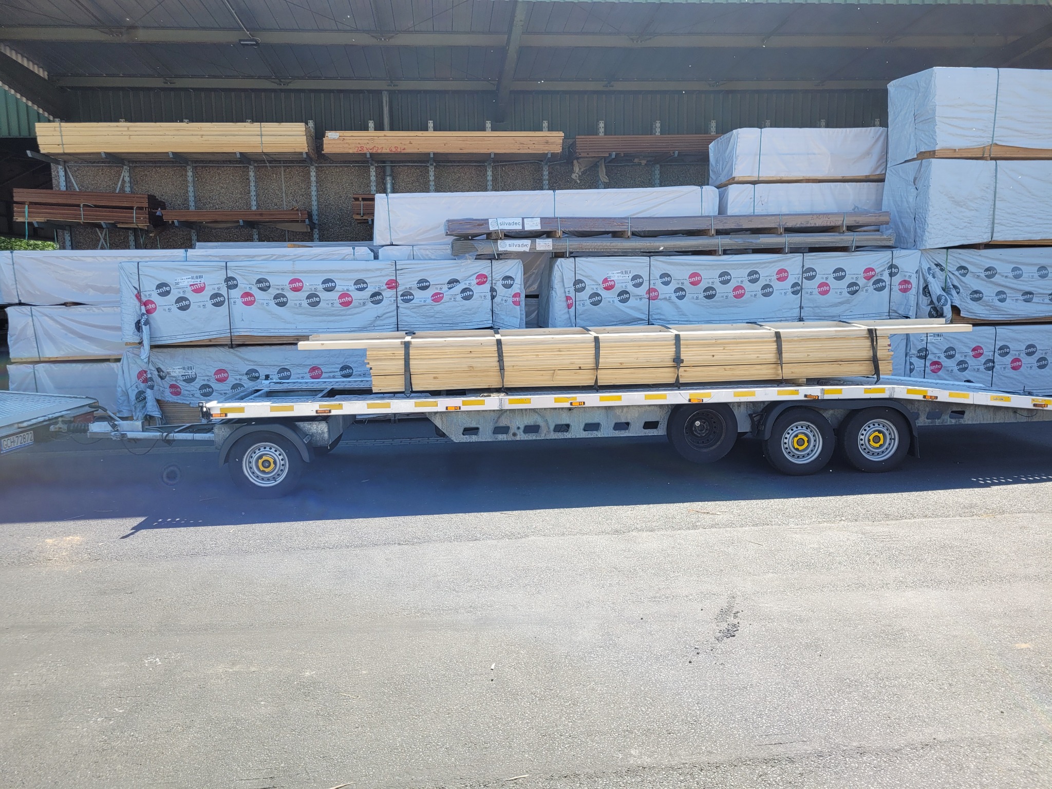 Przyczepa z drewnem budowlanym na tle magazynu z zapasami drewna.