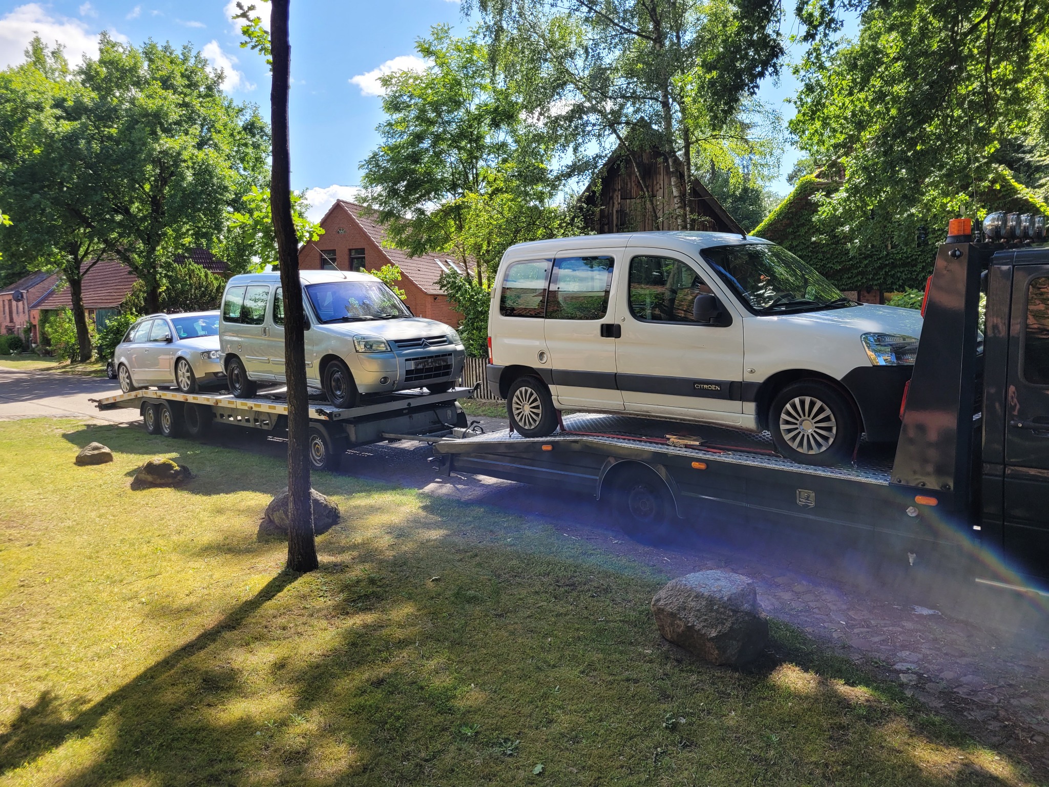 Laweta przewozi trzy samochody, w tym dwa busy, na tle zielonego trawnika i zabudowań mieszkalnych.