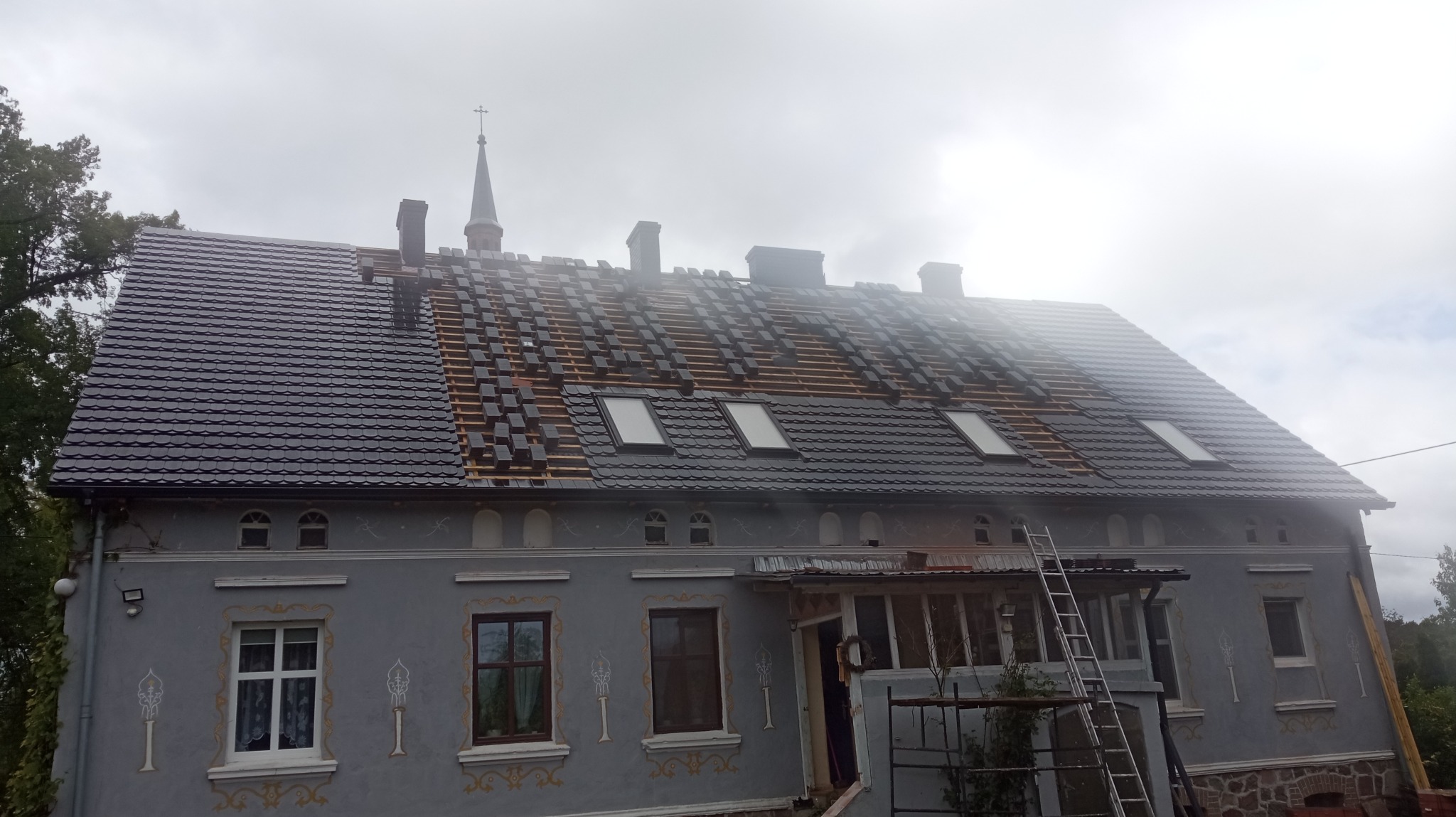 Częściowo pokryty dach z czarną dachówką na szarym budynku z oknami dachowymi. Widoczna drabina i fragmenty konstrukcji dachu. Prace dekarskie w toku.