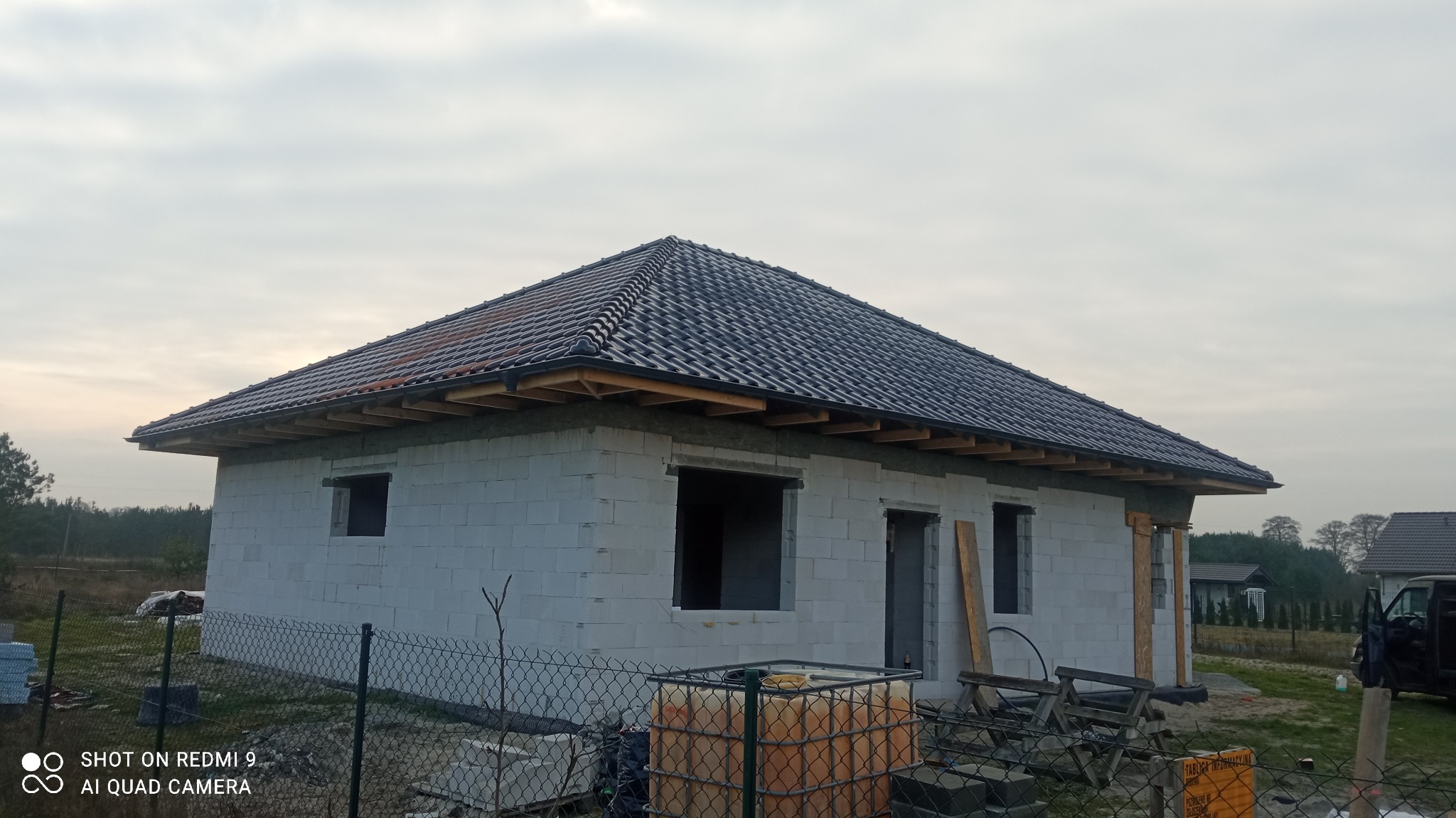 Niedokończony dom z szarego bloczka z częściowo położonym ciemnym dachem z dachówki ceramicznej, widoczne elementy drewniane konstrukcji dachu, otwarte otwory okienne i drzwiowe.