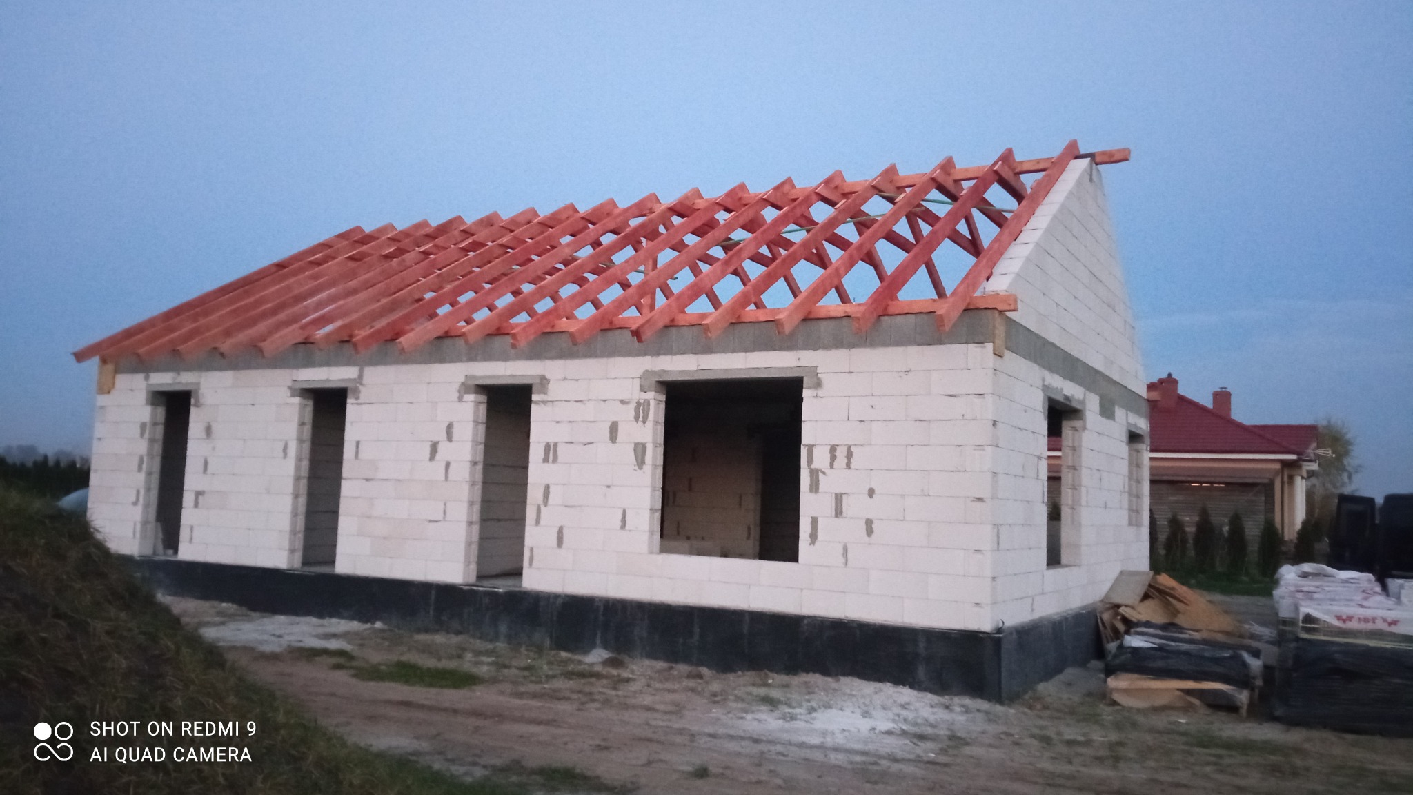 Wznoszony dom jednorodzinny z białych bloczków, z widoczną konstrukcją więźby dachowej z czerwonego drewna, na tle błękitnego nieba o zmierzchu.