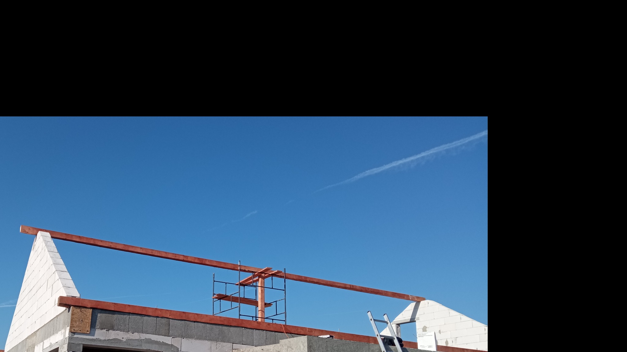 Budowa domu jednorodzinnego: widok na szczyt ściany z zamontowaną drewnianą konstrukcją dachu, rusztowaniem i drabiną na tle błękitnego nieba.