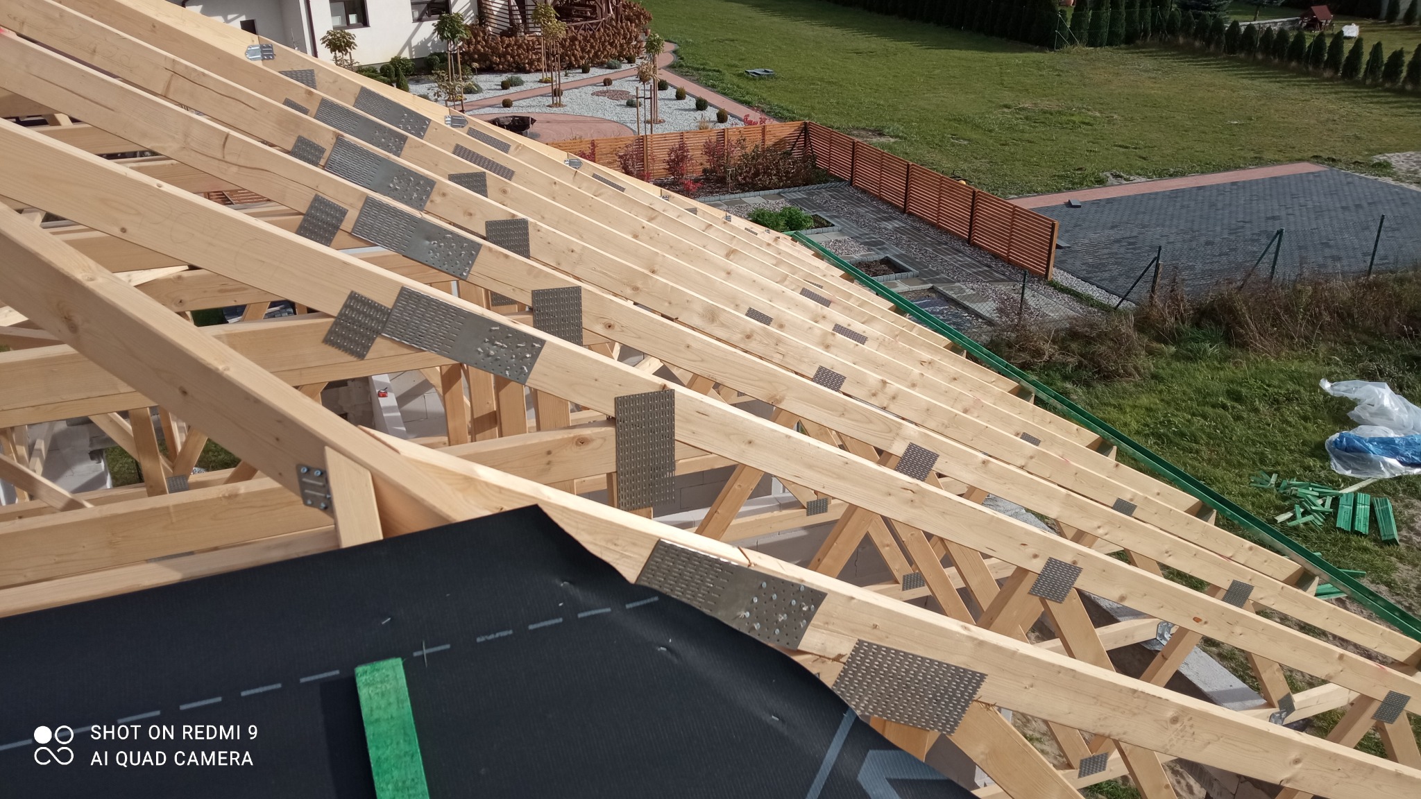 Drewniana konstrukcja dachu z widocznymi metalowymi łącznikami, czarną membraną dachową i elementami obróbek blacharskich, na tle zielonego trawnika i fragmentu domu.