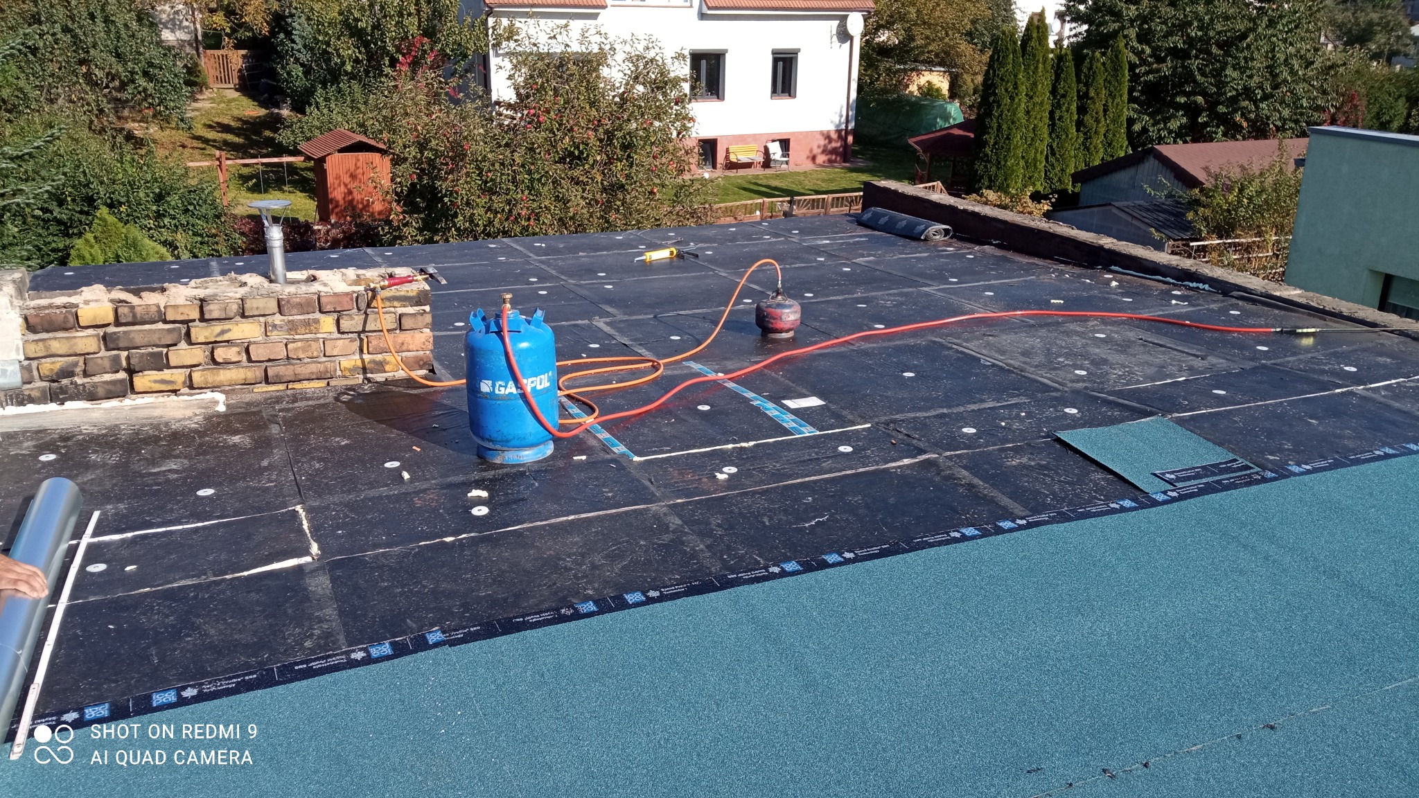 Widok z góry na płaski dach pokryty czarną papą z widocznymi punktami mocowania, na którym stoją butle gazowe, palnik dekarski, fragment komina z cegły oraz rolka papy przygotowana do montażu.