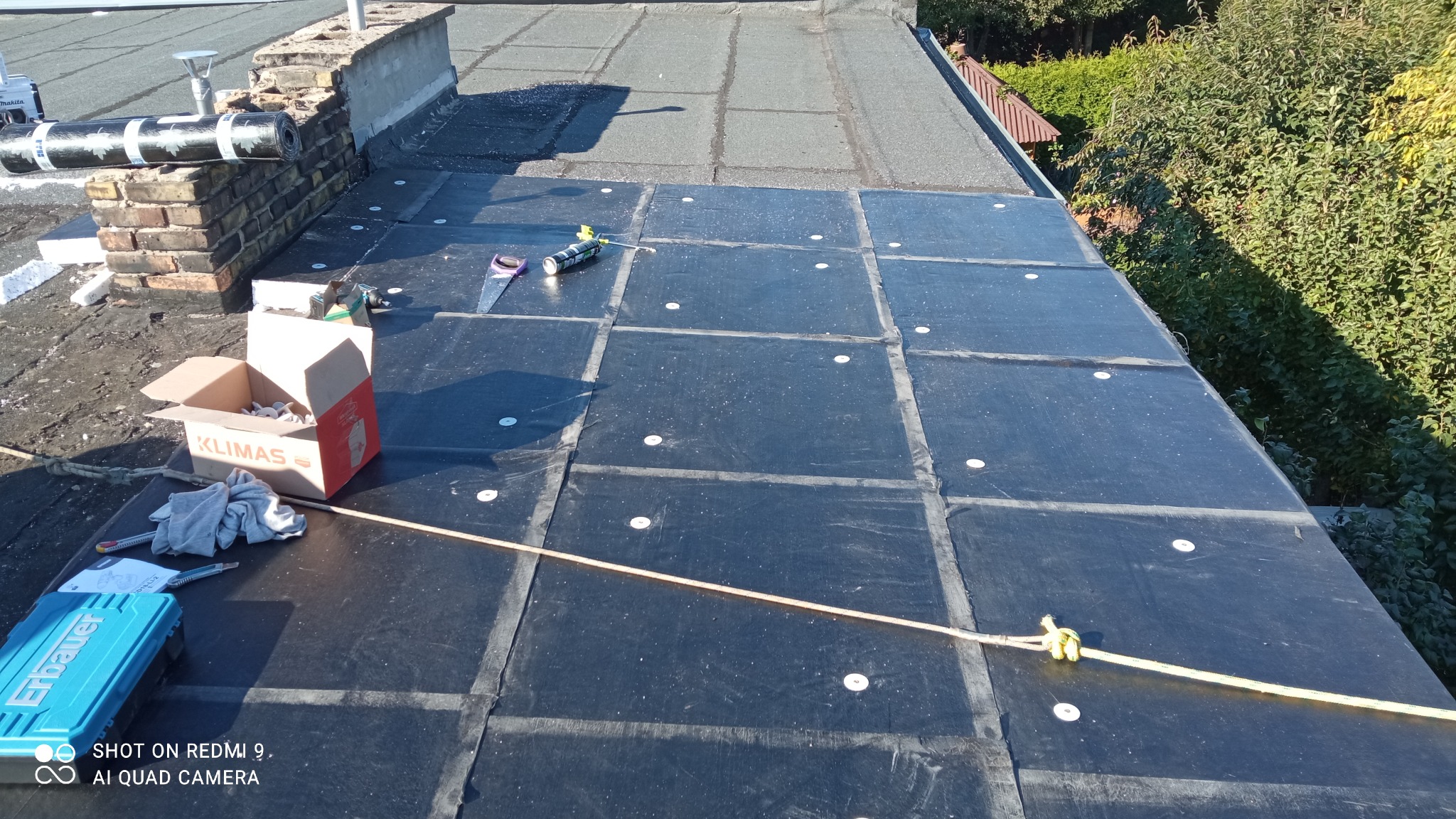 Nowa warstwa hydroizolacyjna z papy termozgrzewalnej, mocowana mechanicznie do dachu płaskiego, z widocznym kominem, narzędziami pracy oraz liną zabezpieczającą.
