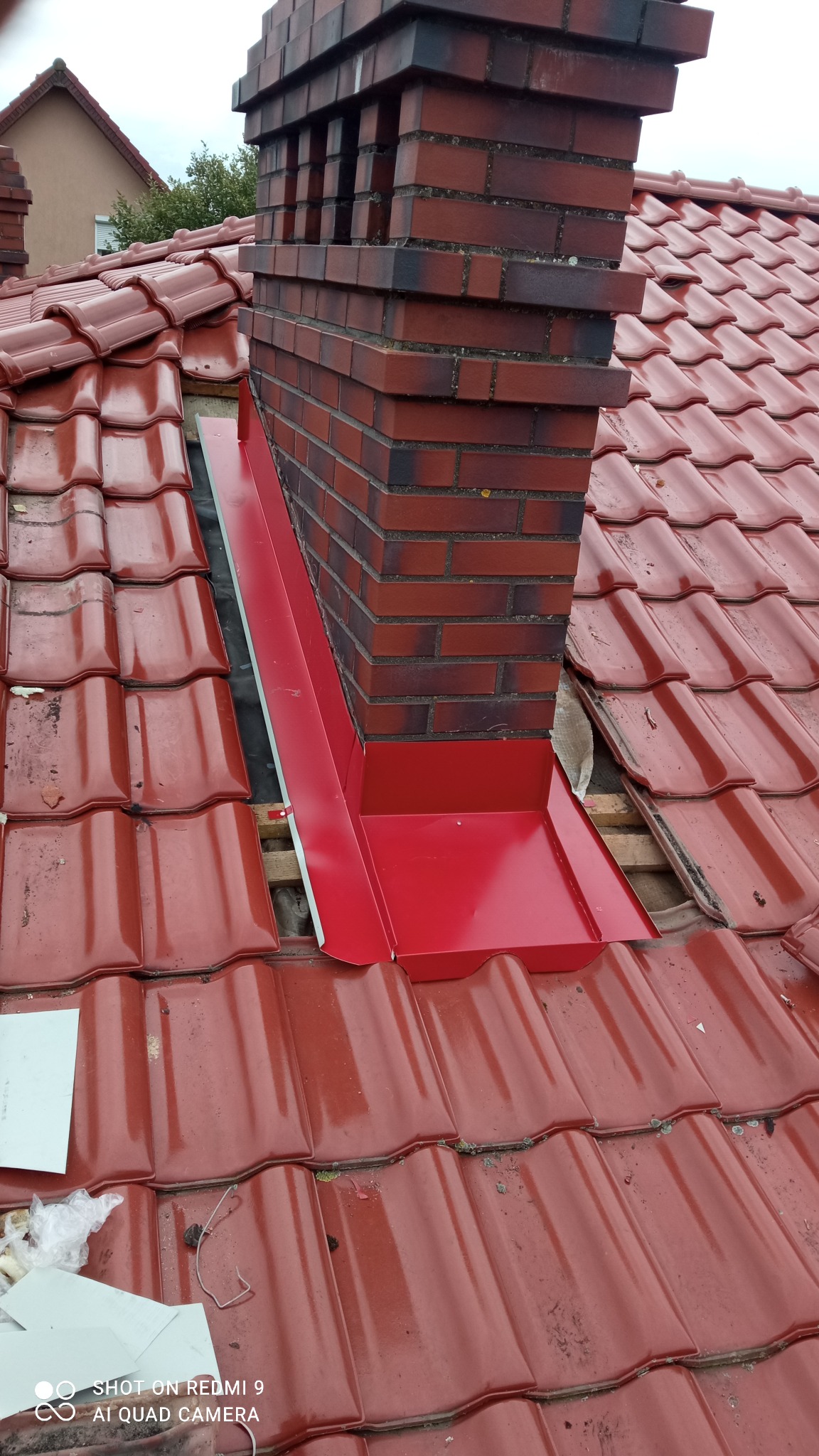 Czerwona dachówka ceramiczna z nową, czerwoną obróbką blacharską komina, widoczne fragmenty konstrukcji dachu, ceglany komin z ciemnymi fugami.