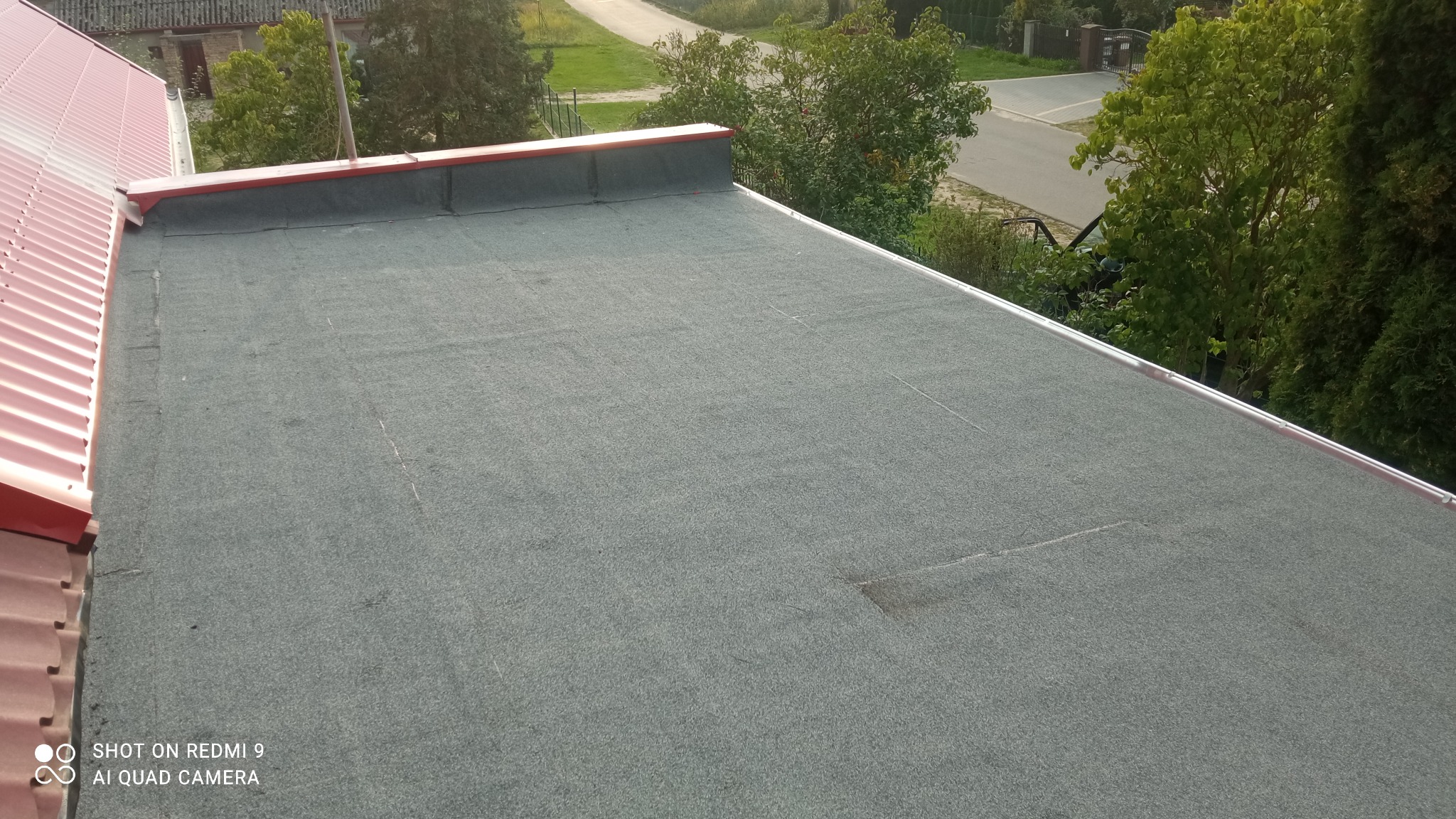 Widok z góry na płaski dach pokryty papą termozgrzewalną z widocznymi łączeniami, z fragmentem dachu skośnego z blachy trapezowej w kolorze czerwonym, w tle zieleń drzew i droga.