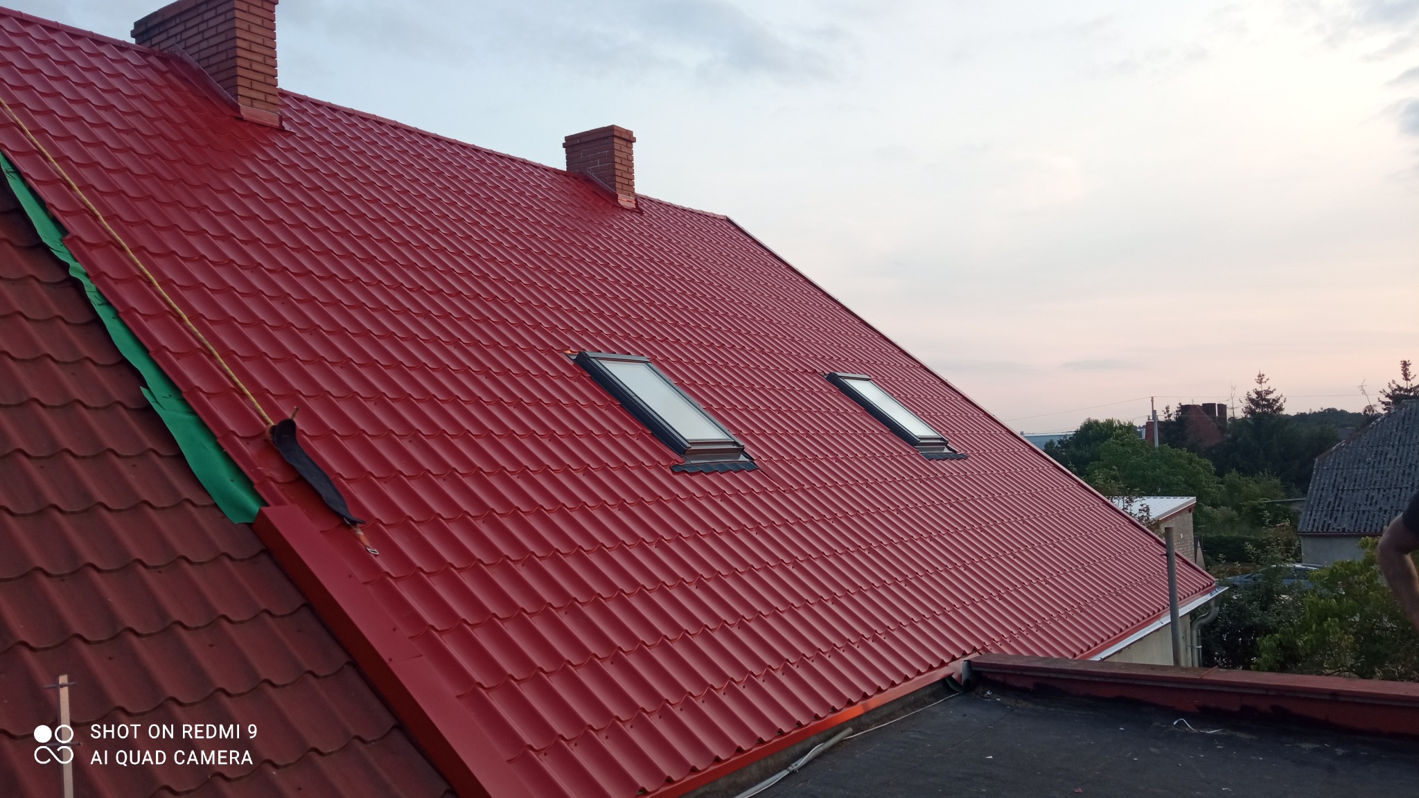 Czerwony dach z blachodachówki z dwoma oknami dachowymi i kominem, widoczne fragmenty zielonej folii zabezpieczającej na krawędzi dachu.