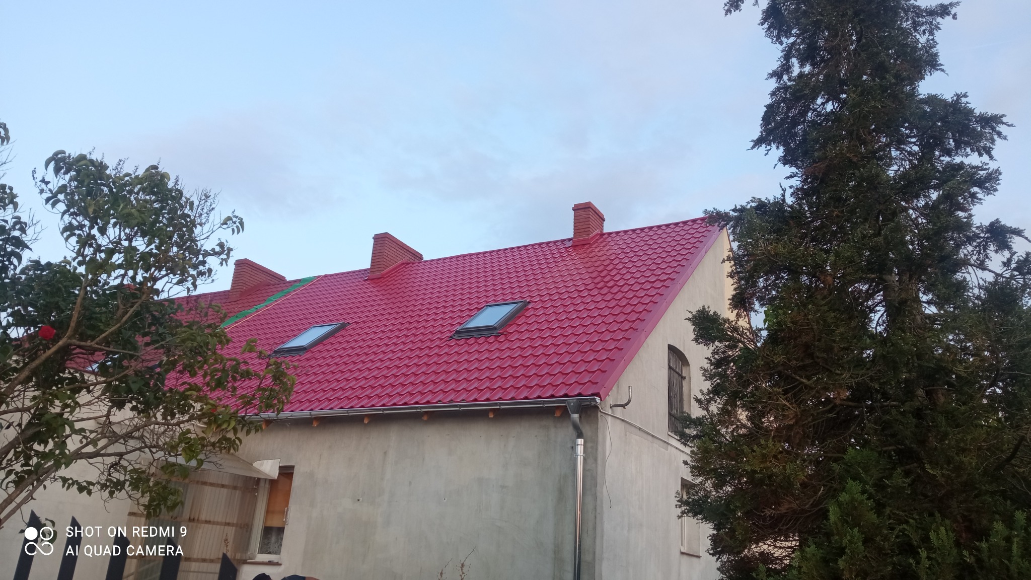 Czerwony dach z dachówką bitumiczną, z dwoma oknami dachowymi, kominami z cegły i nową rynną, na tle niebieskiego nieba, widoczny z poziomu ziemi.