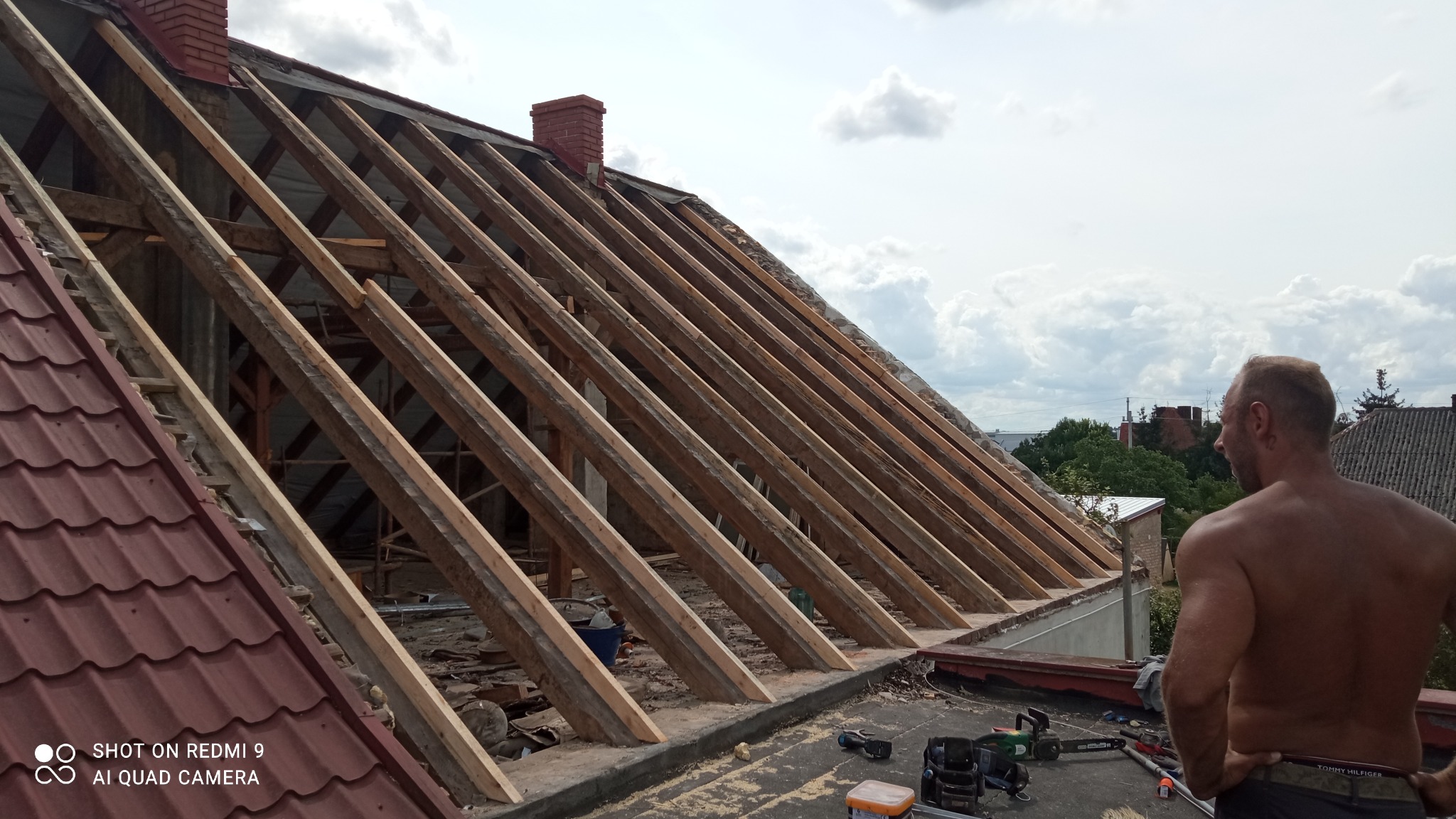 Konstrukcja drewniana dachu z widocznym kominem z czerwonej cegły, częściowo pokryta czerwoną blachodachówką, w tle niebo z chmurami i sylwetka mężczyzny bez koszulki.