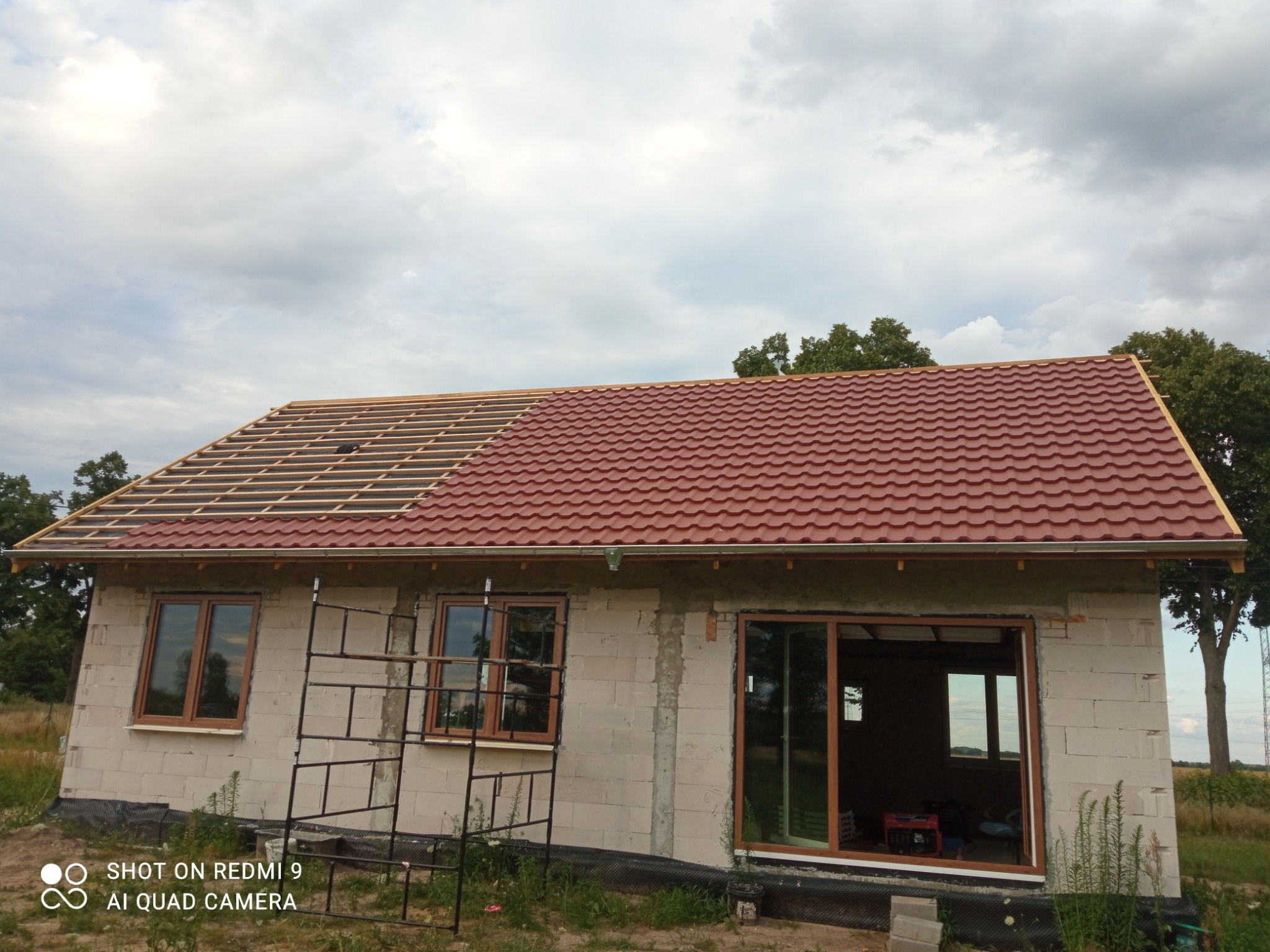 Budynek jednorodzinny w trakcie prac dekarskich, widoczna konstrukcja więźby dachowej z jednej strony i czerwona blachodachówka z drugiej, tło pochmurne niebo.