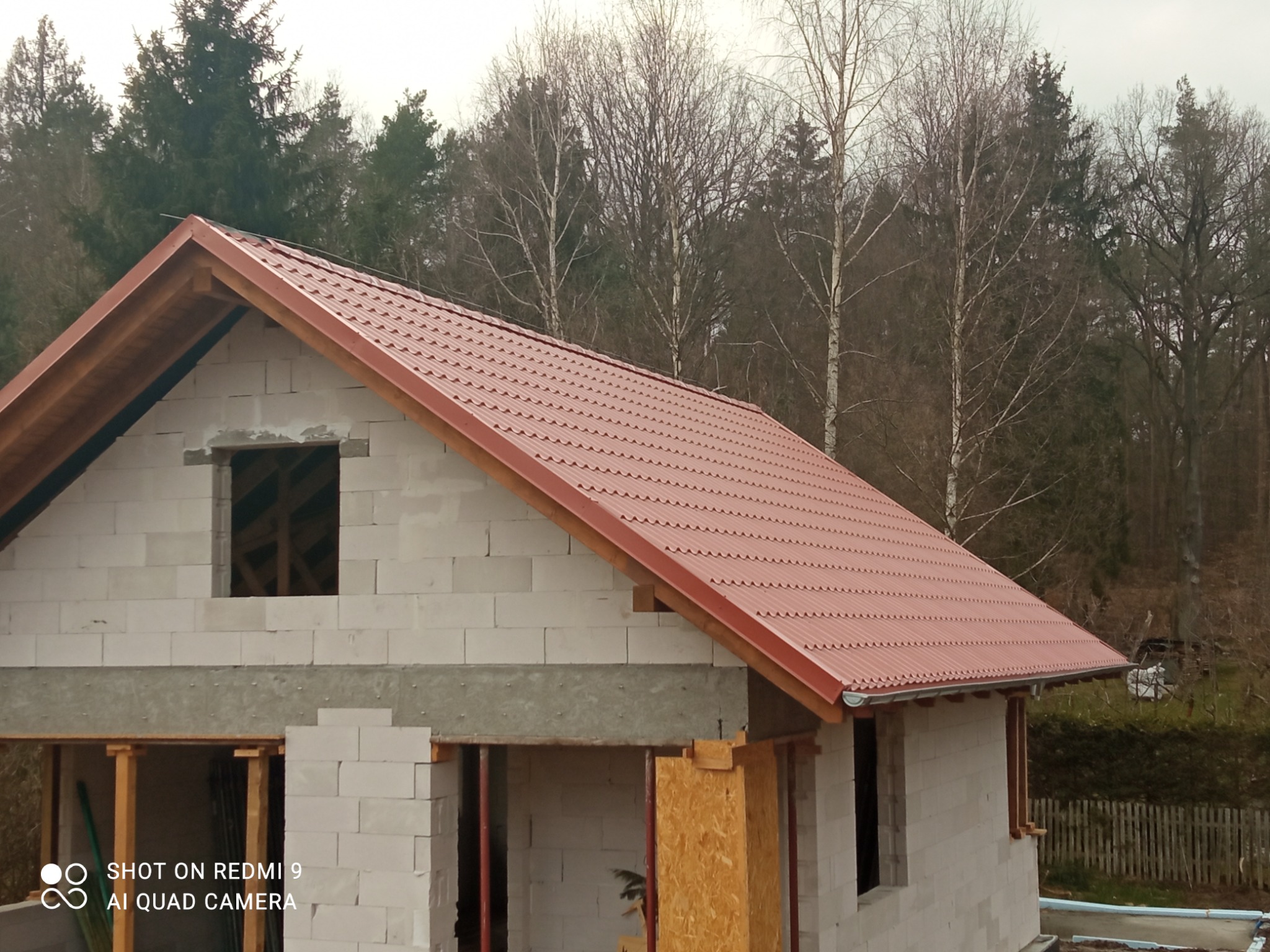 Dom w trakcie budowy z zamontowanym, czerwonym dachem z blachy trapezowej i zamontowanymi rynnami, widok na las w tle.