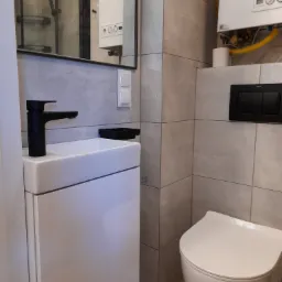 Nowoczesna łazienka z szarymi płytkami, wiszącą toaletą, białą szafką z umywalką i czarną baterią, widoczny bojler gazowy i kabina prysznicowa w lustrze.