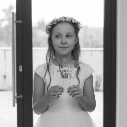 Jakub Kowalski - Portret dziewczynki w białej sukience komunijnej, z wiankiem na głowie, trzymającej dekorację 'Pierwsza Komunia Święta Sary' na tle przeszklonych drzwi.