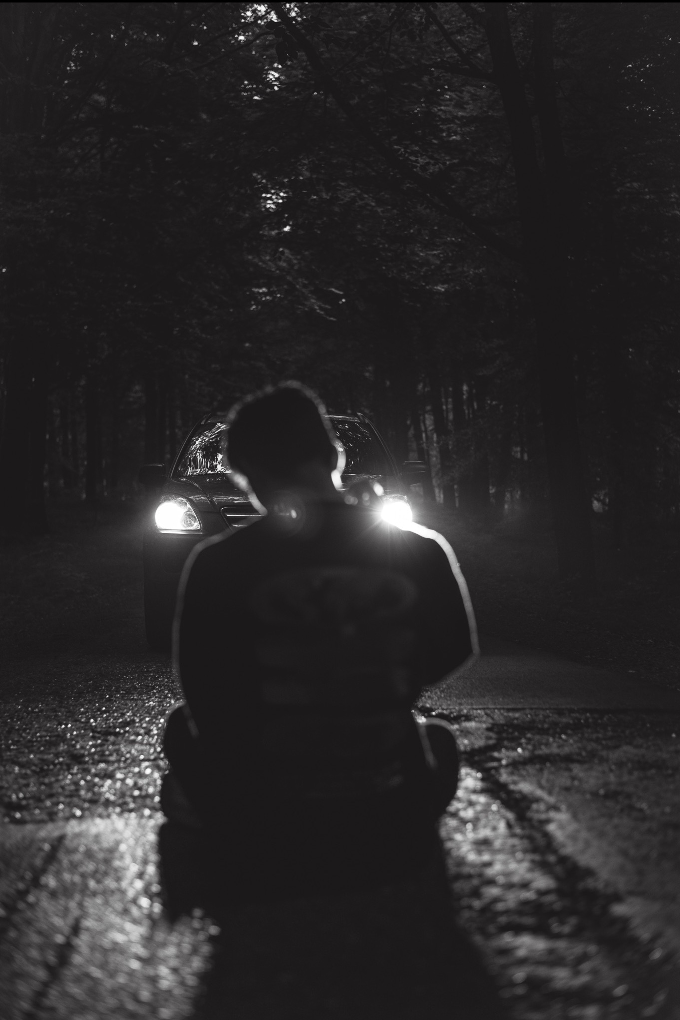 Czarno-białe zdjęcie z tyłu siedzącej osoby na mokrej drodze, z samochodem w tle oświetlonym reflektorami, w otoczeniu drzew.