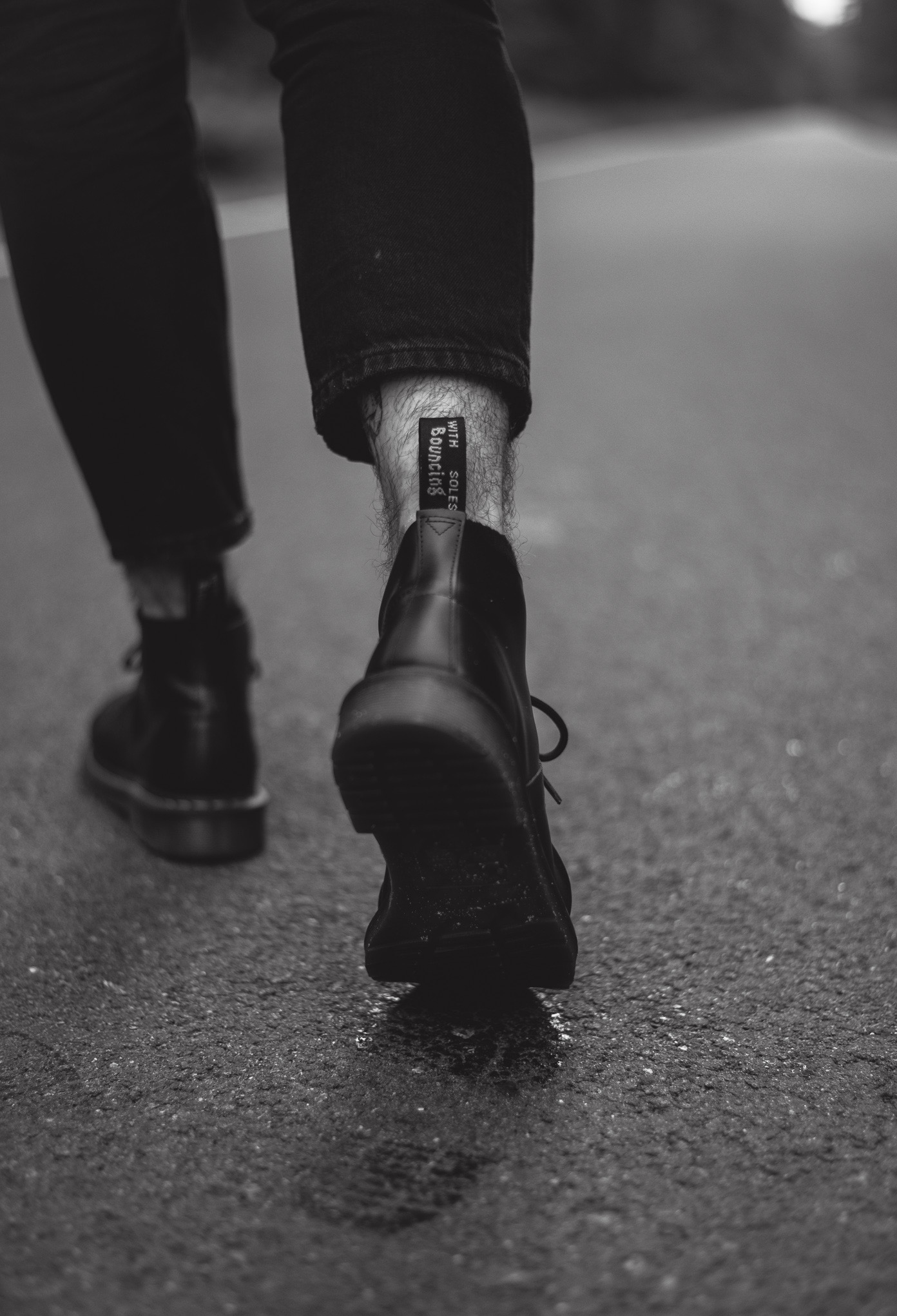 Czarno-białe zbliżenie na czarne buty typu martens na asfalcie, widok z tyłu, z widoczną metką 'WITH BOUNCING SOLES' i fragmentem nogi.