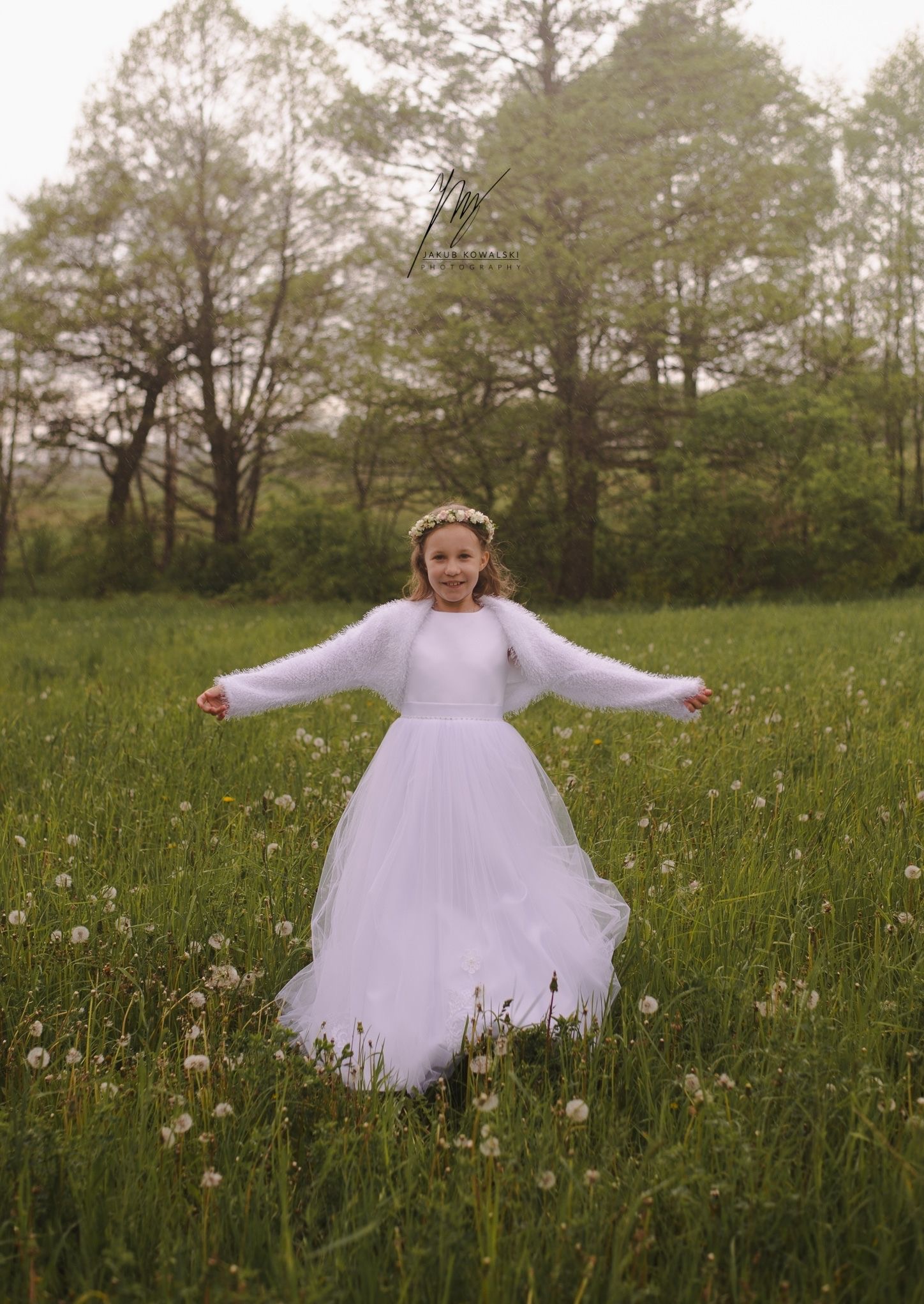 Uśmiechnięta dziewczynka w białej sukience i wianku na głowie, rozkłada ręce na łące z dmuchawcami. W tle drzewa i logo fotografa.