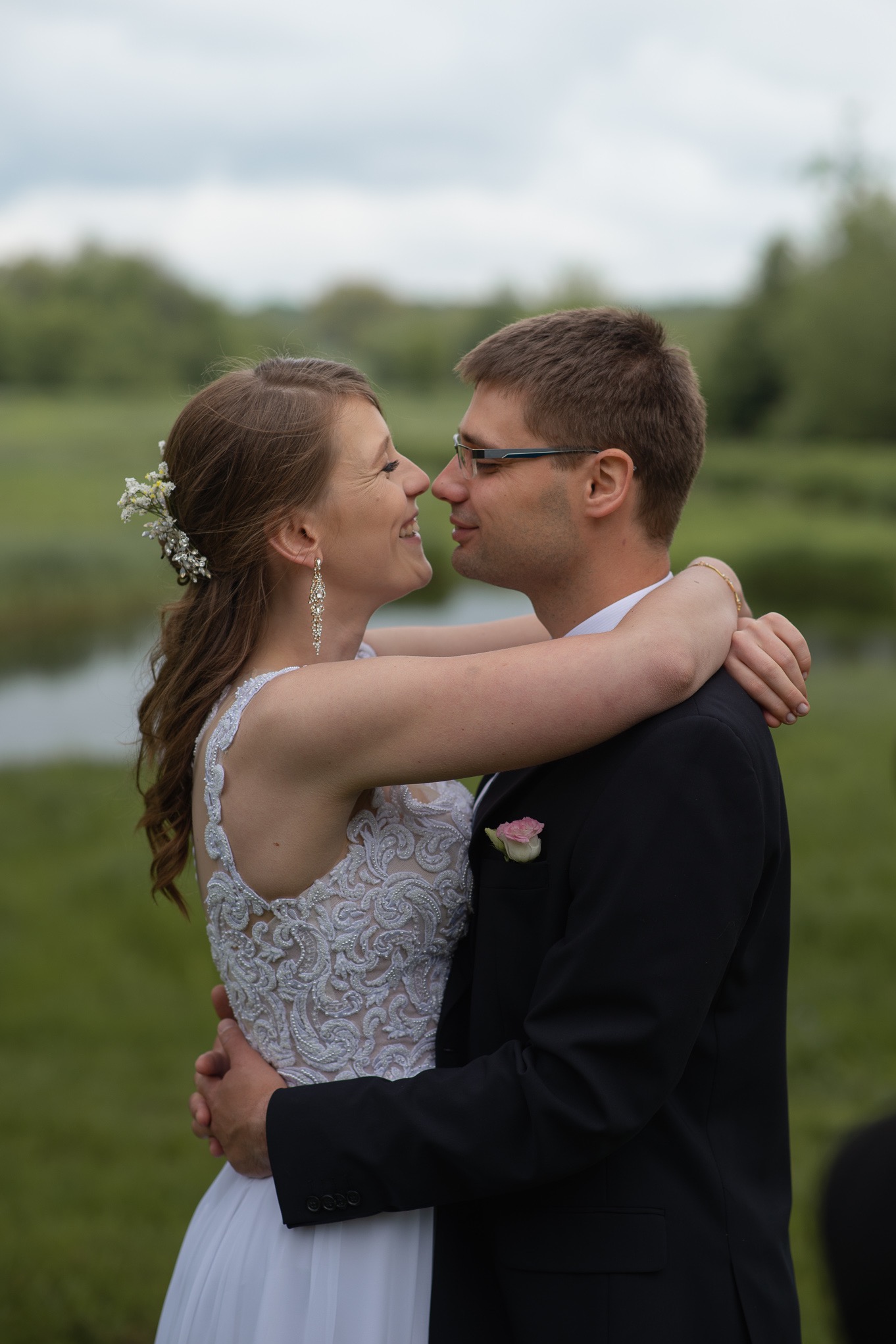 Szczęśliwa para młoda w objęciach, z uśmiechem patrzy na siebie na tle zieleni i stawu. Ona w koronkowej sukni, on w czarnym garniturze i okularach.