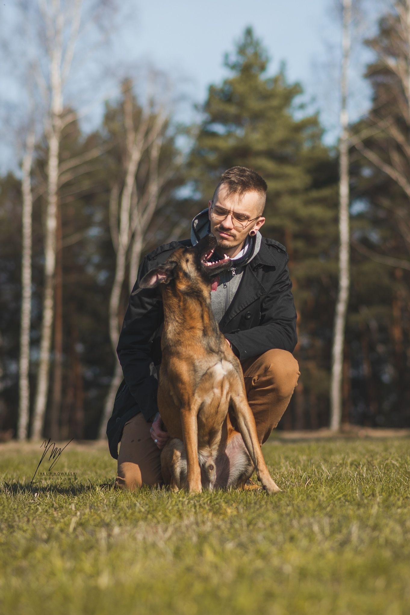Mężczyzna w okularach kuca na trawie, obejmując brązowego psa rasy owczarek belgijski. W tle las z brzozami i drzewami iglastymi w słoneczny dzień.