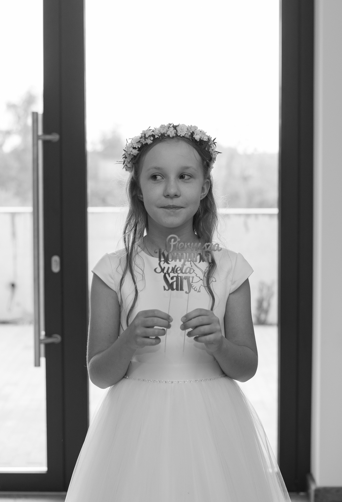 Portret dziewczynki w białej sukience komunijnej, z wiankiem na głowie, trzymającej dekorację 'Pierwsza Komunia Święta Sary' na tle przeszklonych drzwi.