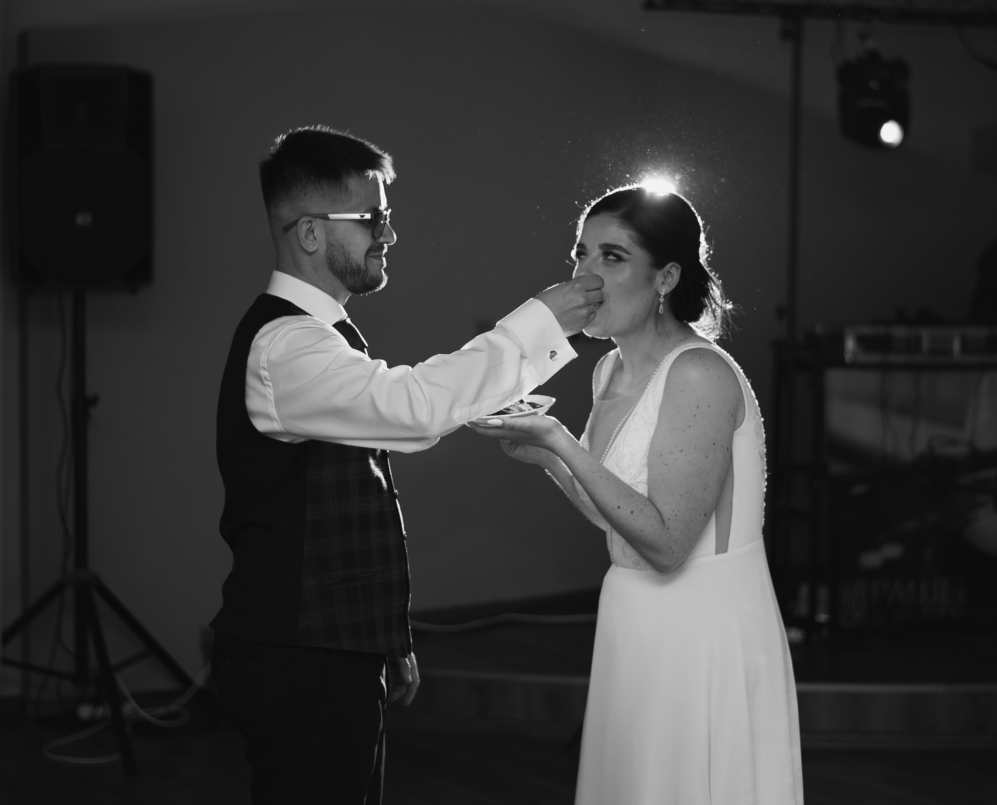 Czarno-białe zdjęcie pary młodej podczas karmienia się tortem na weselu. Pan młody w okularach podaje kawałek tortu pannie młodej w sukni z koronką.