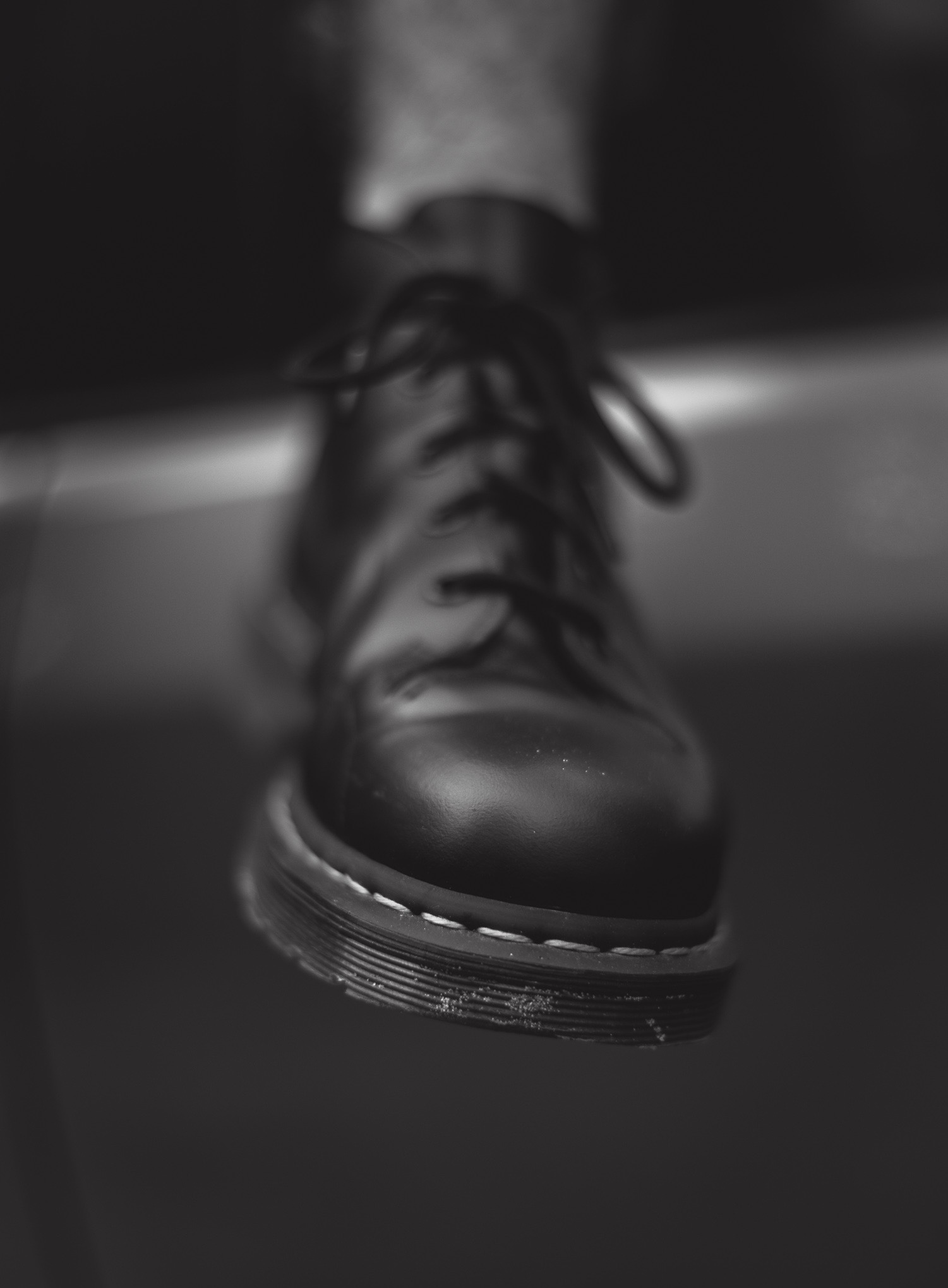 Czarno-białe zbliżenie na czarny, skórzany but z sznurowadłami, ustawiony pod kątem. Widoczna charakterystyczna podeszwa z białym przeszyciem i detale skóry.