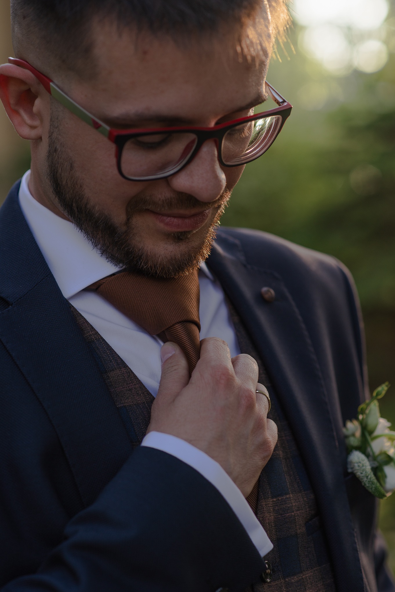 Elegancki mężczyzna w garniturze, poprawiający krawat. Widoczny pierścionek na palcu, butonierka z białych kwiatów. Portret w miękkim świetle, z zamazanym tłem.