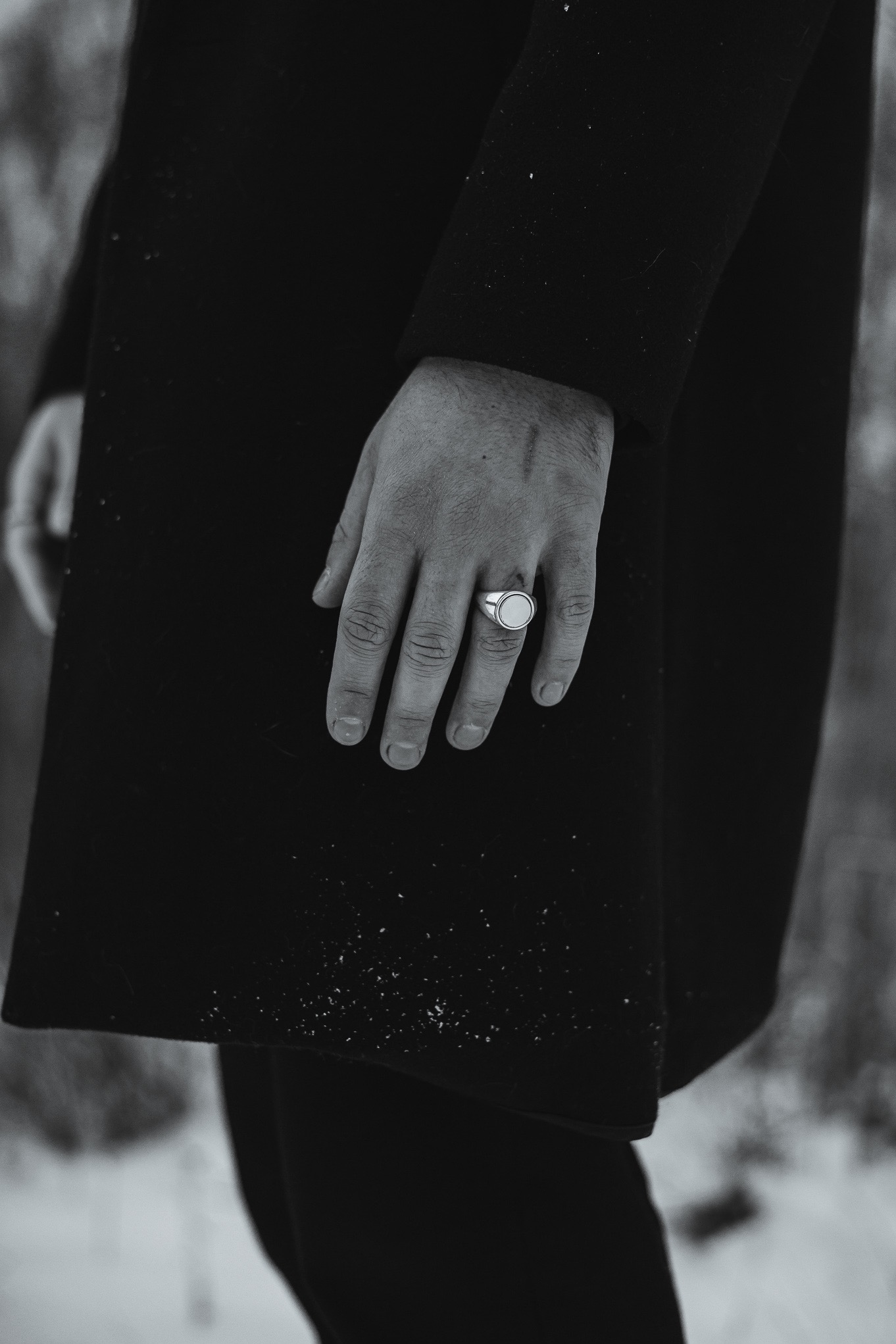 Czarno-białe zbliżenie dłoni mężczyzny z sygnetem na tle ciemnego płaszcza i zimowego krajobrazu. Delikatne ziarno dodaje tekstury.