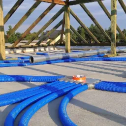 Widok z góry na niebieskie, karbowane rury wentylacyjne rozchodzące się promieniście z metalowej skrzynki rozdzielczej, zabezpieczonej pomarańczową taśmą, ułożone na szarej powierzchni...