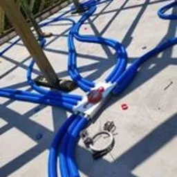 Rozprowadzenie niebieskich rur instalacji wodnej na podłodze, widoczna metalowa skrzynka rozdzielcza z czerwonym zaworem, fragment drewnianej konstrukcji dachu w tle.