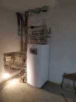 Nowy, biały piec centralnego ogrzewania zamontowany w rogu pomieszczenia, z widocznymi podłączeniami rur miedzianych i plastikowych. Ściany w stanie surowym, widoczne ślady po kuciu. Na podłodze...