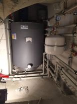 Szary, pionowy zbiornik na wodę w piwnicy, z białym zbiornikiem wyrównawczym obok, rury instalacji grzewczej na ścianie, widoczne manometry i zawory.