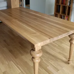 Stół z jasnego drewna o gładkim blacie i toczonych nogach, ustawiony na drewnianej podłodze w jasnym wnętrzu z widoczną biblioteczką w tle.