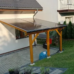 Drewniana wiata przy ścianie domu z przezroczystym zadaszeniem, widoczne słupy z żółtego drewna i fragment kostki brukowej oraz trawnika.